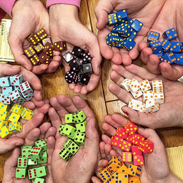 A group of hands displaying colorful Tenzi dice in various colors, including yellow, green, blue, black, and orange. The dice are arranged in a way that highlights their vibrant designs, gathering together in a fun and lively game setting.