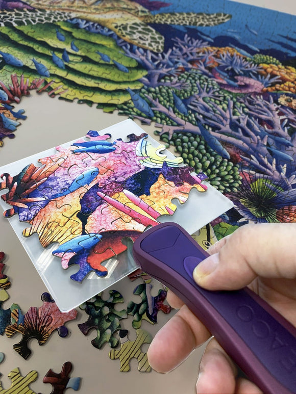 A person uses The Puzzle Scoop, a jigsaw puzzle tool, to lift and place a bright, colorful puzzle piece depicting fish and coral. The background shows an underwater scene with various shades of green and blue, featuring a turtle and vibrant corals.