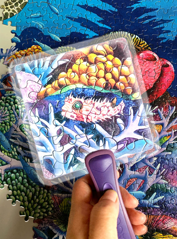 A colorful jigsaw puzzle titled 'The Puzzle Scoop' by CEACO is partially assembled, featuring a vibrant underwater scene with coral and aquatic life. A hand holds a transparent puzzle scoop above a section of the puzzle, helping to navigate the pieces. The colors include shades of blue, green, and various coral colors, creating a visually engaging image that's perfect for puzzle enthusiasts.