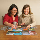 Two women sitting at a wooden table, engaged in assembling the jigsaw puzzle titled 'Where to Next?' by Cobble Hill. One woman, wearing a red shirt and glasses, is picking up a puzzle piece, while the other, dressed in a cozy beige sweater, is smiling and looking at the puzzle. A steaming mug sits on the table beside them, and scattered puzzle pieces can be seen around. The puzzle features vibrant illustrations of famous landmarks and travel destinations.
