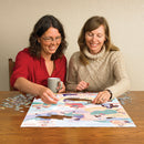 Two women assembling the Literary Women jigsaw puzzle by Cobble Hill at a wooden table. One woman holds a mug while placing a puzzle piece, and the colorful puzzle features illustrations of famous female literary figures.