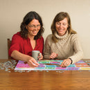 Two women enjoying a jigsaw puzzle titled 'Papel Picado' by Cobble Hill, working together at a wooden table with a colorful puzzle featuring intricate paper cut designs scattered around them. One woman holds a coffee cup while the other focuses on placing a piece.