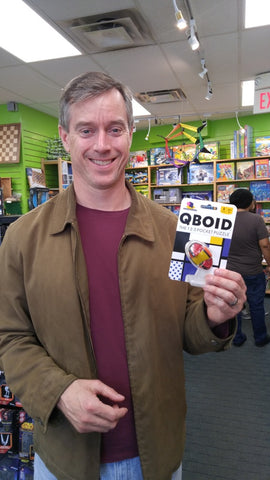 A man smiling in a store holding the Qboid mechanical puzzle by Brainwright. The product is displayed in its packaging, featuring vibrant colors and a unique design. The background shows shelves filled with various games and puzzles, highlighting a playful atmosphere.