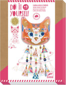 The Kitty Carillons Wind Chimes DIY kit from DJECO features a vibrant cat face design adorned with colorful patterns and glitter. The wind chime includes decorative elements like geometric shapes and playful beads, perfect for kids aged 5 and up to create a whimsical outdoor decor.