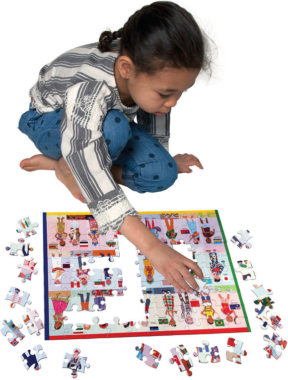 A child concentrating on assembling the 100-piece 'Children of the World' jigsaw puzzle by Eeboo. The puzzle features colorful illustrations of children from various cultures, with several pieces scattered around her. The child wears a striped shirt and blue polka dot pants and is kneeling on the floor, focused on fitting a puzzle piece into place.