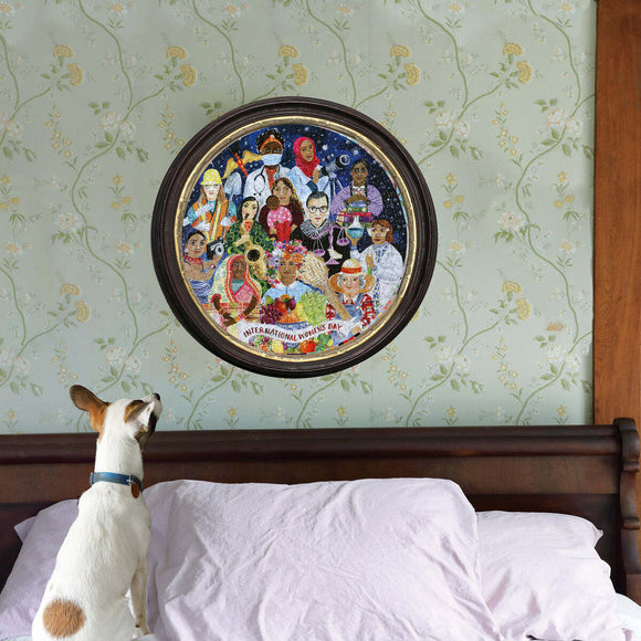 The International Women's Day jigsaw puzzle by Eeboo features a colorful and vibrant artwork depicting diverse women celebrating their achievements. The puzzle is displayed in a round frame on a wall with floral wallpaper, while a curious dog looks up at it from a bed adorned with a light purple blanket. The scene captures a cozy, inviting atmosphere for puzzle enthusiasts.