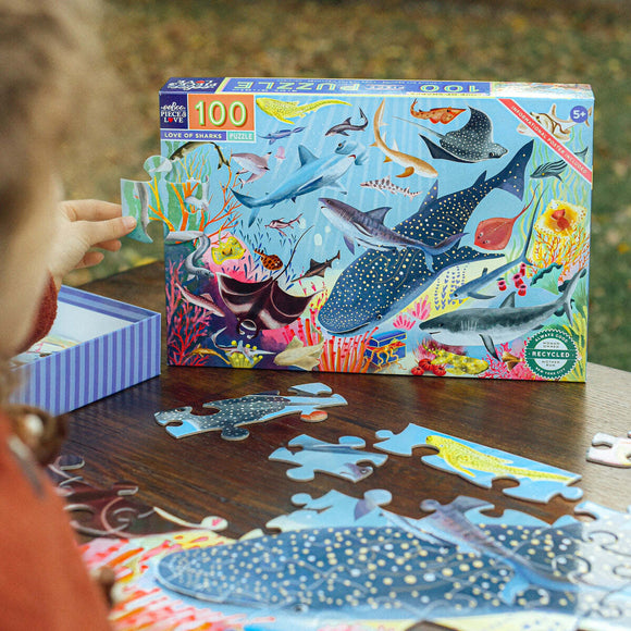 The Love of Sharks jigsaw puzzle by Eeboo features vibrant illustrations of various shark species and marine life. The colorful box shows a scene of sharks amidst coral and underwater plants. A child is assembling a piece of the puzzle on a wooden table, with several puzzle pieces scattered around.