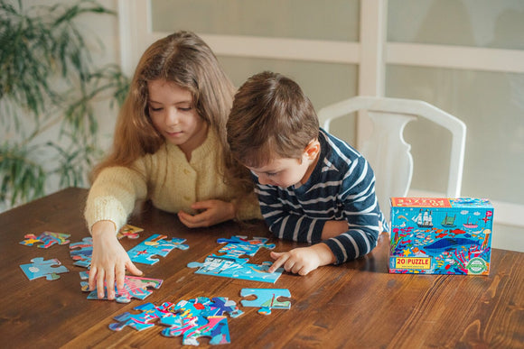 Two children engaged in a jigsaw puzzle activity on a wooden table. The girl, with long hair, is placing a puzzle piece while the boy in striped attire looks at another piece. The colorful box of the 20-piece Sea Exploration jigsaw puzzle by Eeboo is visible nearby, showcasing vibrant ocean-themed illustrations.
