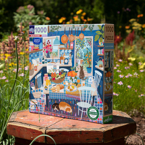 A colorful 1000-piece jigsaw puzzle titled 'Blue Kitchen' by Eeboo. The image features a vibrant kitchen scene with blue decor, a dining table laden with fruits and baked goods, and playful cats lounging on the floor. This intricately illustrated puzzle is perfect for family fun and relaxation.