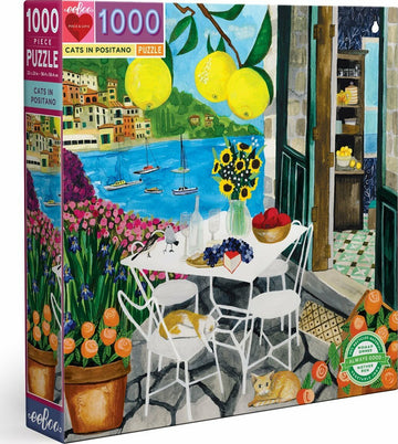 Eeboo's Cats in Positano 1000-piece jigsaw puzzle featuring a colorful illustration of a seaside terrace with cats lounging, vibrant flowers, a table set with fruits, and a scenic view of boats on blue waters. The scene captures the essence of Positano with lemon trees and charming architecture in the background.
