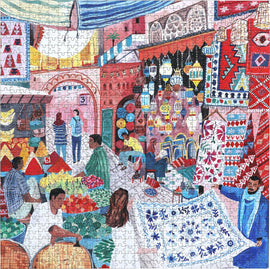 A colorful jigsaw puzzle titled 'Marrakesh' by Eeboo, depicting a vibrant Moroccan market scene. It features various vendors selling fruits, vegetables, and textiles, with traditional lanterns and patterned rugs displayed in the background. The artwork is richly detailed, showcasing a lively atmosphere filled with people interacting and shopping.