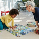 A young child and an adult woman are sitting on the floor, engaged in assembling a colorful jigsaw puzzle titled 'Within the Biome' by Eeboo. The child, wearing a bright yellow sweater and blue pants, is focused on placing a puzzle piece into a vibrant scene featuring various plants and animals. The adult, with silver hair tied back, watches with a smile, offering support. A box containing the puzzle can be seen in the background.