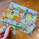 An image of a partially completed jigsaw puzzle titled "Within the City" by Eeboo, featuring colorful urban scenes with buildings, vehicles, and people. A player is placing a piece on the puzzle, which consists of 48 large pieces.