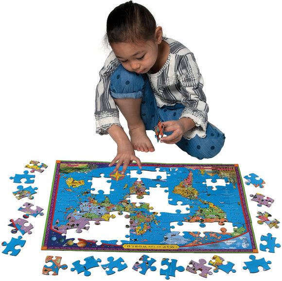 A young child assembling a colorful 100-piece jigsaw puzzle of a world map. The puzzle features vivid illustrations of continents, landmarks, and animals, with several pieces scattered around. The child, wearing a striped shirt and denim shorts, focuses intently on fitting a piece into the puzzle.