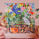 A vibrant jigsaw puzzle titled 'Wild Things' by Eeboo, showcasing a colorful scene filled with wildflowers, animals like a rabbit and squirrels, and butterflies. A child's hands are seen working on the puzzle, placing a piece into the lively illustration.