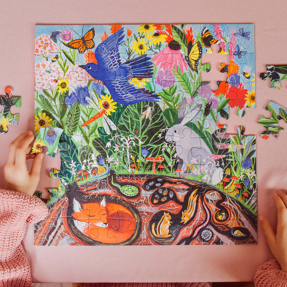 A vibrant jigsaw puzzle titled 'Wild Things' by Eeboo, showcasing a colorful scene filled with wildflowers, animals like a rabbit and squirrels, and butterflies. A child's hands are seen working on the puzzle, placing a piece into the lively illustration.