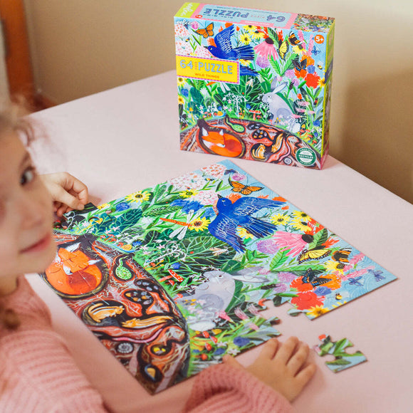 A young child is working on the 'Wild Things' jigsaw puzzle by Eeboo, with colorful pieces featuring animals and vibrant flora. The puzzle shows intricate designs of various creatures surrounded by vivid flowers and greenery. In the background, the puzzle box is displayed, showcasing the playful artwork. The scene emphasizes creativity and fun in a cozy setting.
