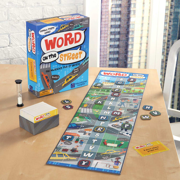 A colorful board game titled 'Word On The Street' by Educational Insights is displayed on a wooden table. The game box features vibrant artwork with cars and text promoting it as a family/party game. The game board is spread out beside the box, showcasing a layout with various letters, words, and graphics of city scenes. An hourglass timer and a stack of game cards are also present, indicating gameplay elements and a fun interactive experience.