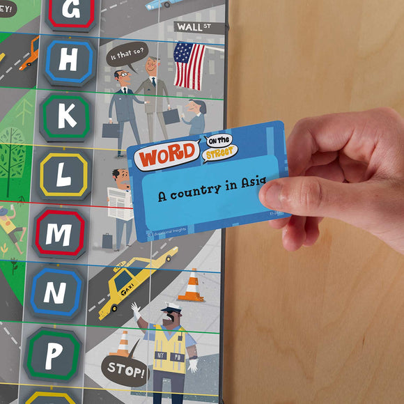 A hand holds a blue 'Word On The Street' game card that reads 'A country in Asia'. The game board features colorful graphics, including street signs, characters, and vehicles, set against a city backdrop.