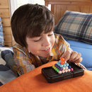 A young boy with brown hair is engaged in playing Kanoodle, a mechanical puzzle by Educational Insights. He is smiling while concentrating on stacking colorful, interlocking pieces on a compact black game board, surrounded by a cozy bedroom setting with blue and plaid bedding.
