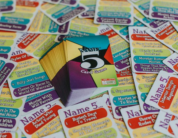 A colorful pack of the Name 5 Card Game by Goliath, surrounded by various game cards featuring prompts like 'Billy Joel Songs' and 'Monster Movies'. The game encourages players to think quickly and come up with five answers in engaging categories for fun and laughter.