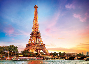 Eurographics jigsaw puzzle featuring a vibrant depiction of the Eiffel Tower in Paris, illuminated against a colorful sunset sky. The puzzle showcases intricate details of the iconic landmark surrounded by lush greenery and the Seine River, perfect for fans of travel and architecture.