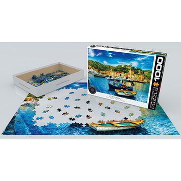A 1000-piece jigsaw puzzle titled 'Portofino Italy' by Eurographics is partially assembled on a flat surface. The puzzle features a colorful image of boats docked in a picturesque harbor surrounded by vibrant buildings and lush greenery. The puzzle box is open beside it, showing the remaining puzzle pieces inside.
