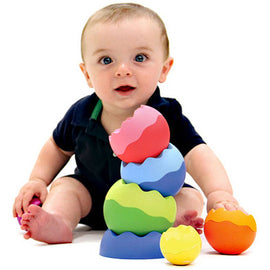 A joyful baby sitting on the floor, engaged with Tobbles - Neo by Fat Brain Toys. The baby is surrounded by colorful, stackable spheres in various sizes, featuring wavy edges and vibrant colors. The child is reaching for a bright orange ball while a stacked arrangement of green, blue, pink, and purple spheres is in front of them, showcasing the educational and playful aspects of this toy designed for developing motor skills and hand-eye coordination.