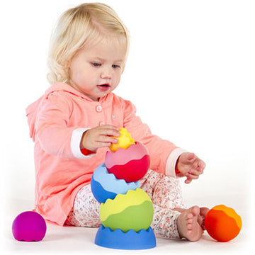 A young child plays with Tobbles - Neo by Fat Brain Toys, stacking colorful, soft, textured shapes in various sizes. The child, dressed in a light pink hoodie, focuses intently on balancing the pieces, which include vibrant spherical and wavy-edged shapes in shades of blue, green, pink, and orange. The play area is bright and inviting, showcasing the engaging and educational nature of this stacking toy.