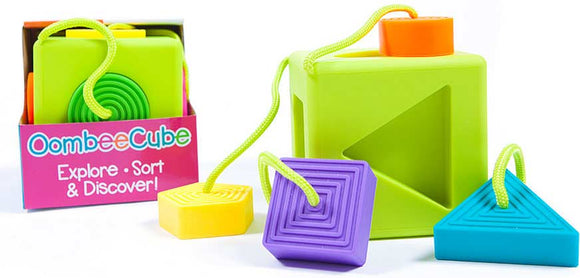 OombeeCube by Fat Brain Toys, featuring colorful geometric shapes in bright green, yellow, purple, and blue. The cube has cutout shapes for sorting and includes textured blocks, encouraging tactile exploration and color recognition for young children. The packaging displays the title 'OombeeCube' with a tagline that invites users to 'Explore, Sort & Discover!'