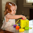 A young child engages with the OombeeCube by Fat Brain Toys, a colorful interactive game designed for early learning. The child, with a bow in her hair, is placing a colorful shape into the cube while sitting at a table. The OombeeCube features various shapes and vibrant colors to enhance sensory and cognitive development.