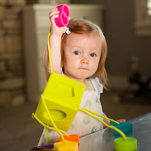 A young child playing with the OombeeCube from Fat Brain Toys. The child holds a pink shape while interacting with a vibrant yellow cube. The cube features cut-out shapes and colorful connectors, promoting hands-on learning and motor skill development.