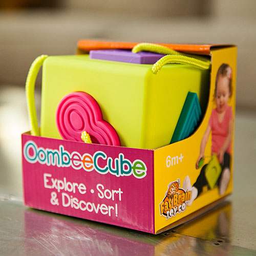 The OombeeCube by Fat Brain Toys is a vibrant, colorful sorting and discovery game designed for children aged 6 months and older. The cube features soft, textured shapes in various colors including pink, purple, and green, making it visually engaging. Its sturdy design is perfect for little hands, promoting sensory exploration and fine motor skills. The packaging showcases a young child happily engaging with the toy, emphasizing its playfulness and educational value.