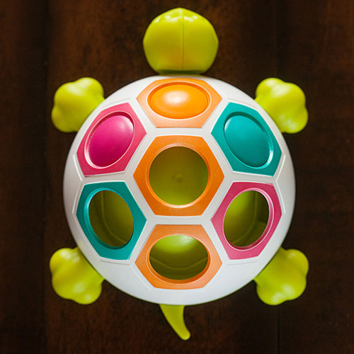 The Pop N Slide Shelly game by Fat Brain Toys features a colorful turtle design with hexagonal slots in vibrant colors including orange, pink, teal, and green. The turtle's body is white with bright green flippers and a tail, presenting an engaging and interactive toy for kids.