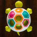 The Pop N Slide Shelly game by Fat Brain Toys features a colorful turtle design with hexagonal slots in vibrant colors including orange, pink, teal, and green. The turtle's body is white with bright green flippers and a tail, presenting an engaging and interactive toy for kids.