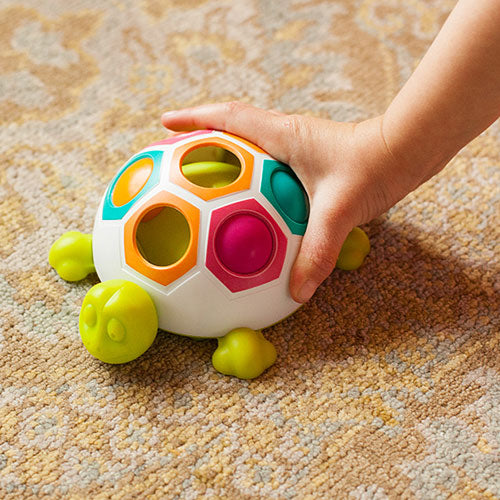 A colorful toy shaped like a turtle featuring hexagonal holes in various bright colors. A child's hand is reaching for the turtle, which has green legs and a smiling face. The background shows a textured carpet, emphasizing the interactive and playful nature of the game for young children.