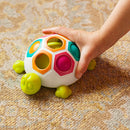 A colorful toy shaped like a turtle featuring hexagonal holes in various bright colors. A child's hand is reaching for the turtle, which has green legs and a smiling face. The background shows a textured carpet, emphasizing the interactive and playful nature of the game for young children.