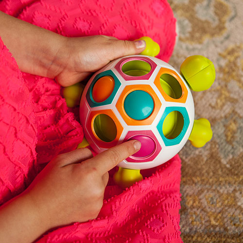A child playing with the Pop N Slide Shelly toy, designed by Fat Brain Toys. The colorful turtle-shaped toy features hexagonal openings and brightly colored buttons that can be pressed, promoting sensory play and fine motor skills. The child is holding the toy in their lap, wearing a pink textured dress, on a patterned rug.
