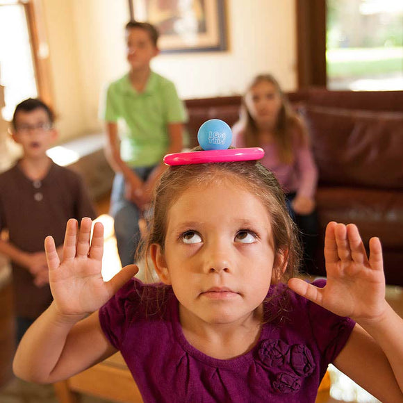 A young girl wearing a purple shirt plays the game 'I Got This' by Fat Brain Toys. She has a blue ball balanced on her head while putting her hands in the air. In the background, three other children observe her with expressions of anticipation.