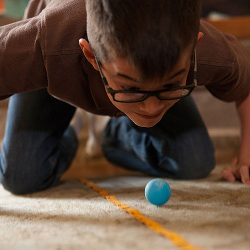 A young boy with glasses is playfully leaning down on a carpeted floor, intently focused on a small blue ball as it rolls along a yellow line. His expression shows excitement and engagement in the game, highlighting a moment of fun and concentration.