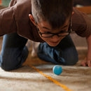 A young boy with glasses is playfully leaning down on a carpeted floor, intently focused on a small blue ball as it rolls along a yellow line. His expression shows excitement and engagement in the game, highlighting a moment of fun and concentration.