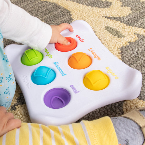 A colorful and engaging sensory toy, the Dimple Duo from Fat Brain features multiple buttons in various shapes and colors, including red triangle, green circle, and yellow oval. A child's hand is pressing the red circle button while sitting on a patterned rug, showcasing the toy's interactive design that promotes tactile play and learning.