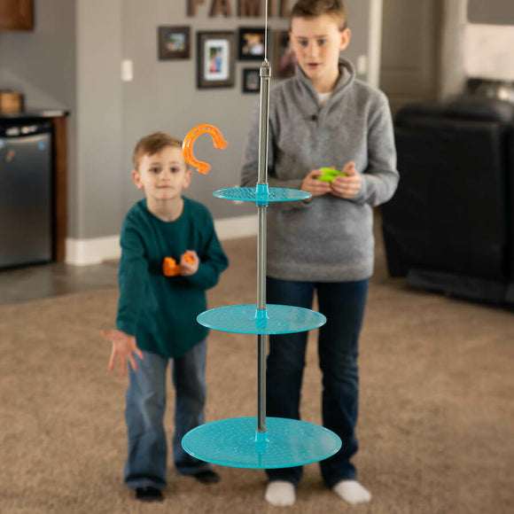Two children play the game 'Swingin' Shoes' by Fat Brain, featuring a multi-tiered structure with hanging blue platforms. One child is reaching to toss an orange hook while the other holds green and orange balls, emphasizing fun and interactive play.