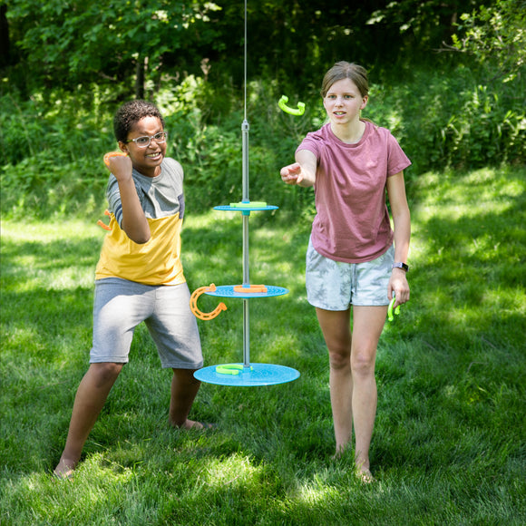 Two children playing with the Swingin' Shoes game in a sunlit green field. One child, wearing a yellow and gray shirt, is in a throwing pose with a smile, while the other, in a pink shirt, is preparing to throw a shoe toward the hanging target. The game features colorful circular platforms and vibrant shoes, encouraging outdoor fun and teamwork.