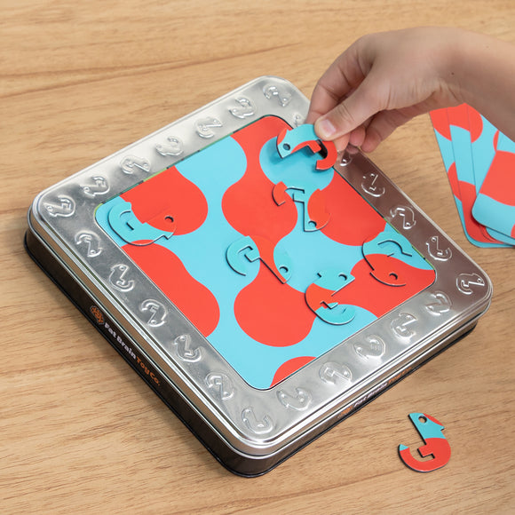 A hand is placing a colorful red and blue puzzle piece into a mechanical puzzle called One in a Chameleon by Fat Brain Toys. The puzzle features a metallic tin with various shaped pieces that fit together in an engaging way, enhancing problem-solving skills.