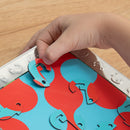 A child's hand is placing a brightly colored, mechanical puzzle piece into a red and blue patterned base. The puzzle features fish-shaped components that fit together, showcasing the engaging and interactive design of the 'One in a Chameleon' mechanical puzzle by Fat Brain Toys. The scene is set on a wooden table surface.