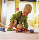 A young child in a green shirt playing on the carpet with colorful wooden blocks and a cheerful toy car featuring a smiling figure. The child is engaged in creative play, exploring shapes and colors.