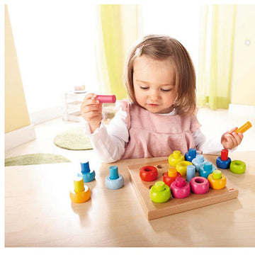 A young child playing with the Pegging Game Rainbow Whirls by Haba. The game features colorful pegs and holes on a wooden board, promoting fine motor skills and color recognition. The child is engaged and holding a pink peg, surrounded by various other colorful pieces.