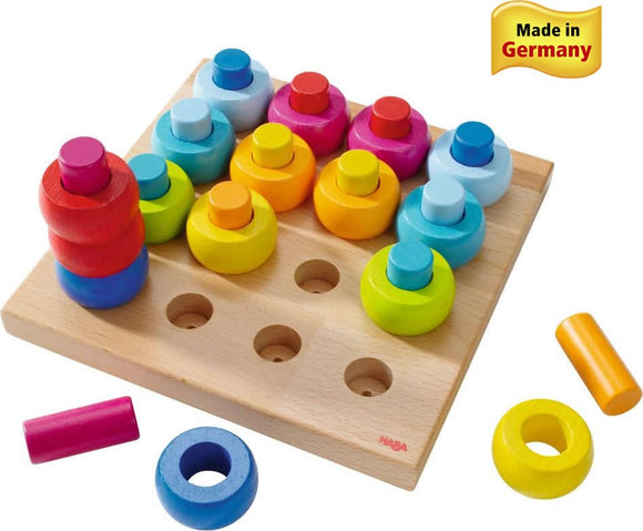 Haba Pegging Game Rainbow Whirls features a wooden board with colorful stacking rings in various shapes and sizes. The game promotes fine motor skills and color recognition, with vibrant hues of red, blue, yellow, green, and pink. Includes both cylindrical and circular pieces, designed for young children, encouraging playful learning. Made in Germany.