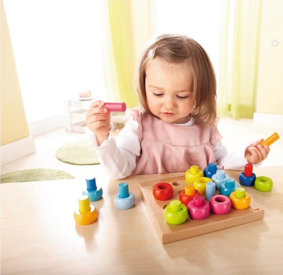 A young girl with brown hair, wearing a pink dress, intently interacts with the Pegging Game Rainbow Whirls by Haba. The game features colorful wooden pieces in various shapes arranged on a light wooden base, enhancing fine motor skills and color recognition.